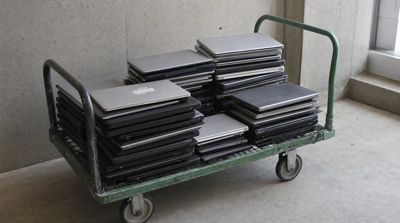 A steel trolley stacked with mismatched retired laptops of different ages and finishes in a plain concrete room.